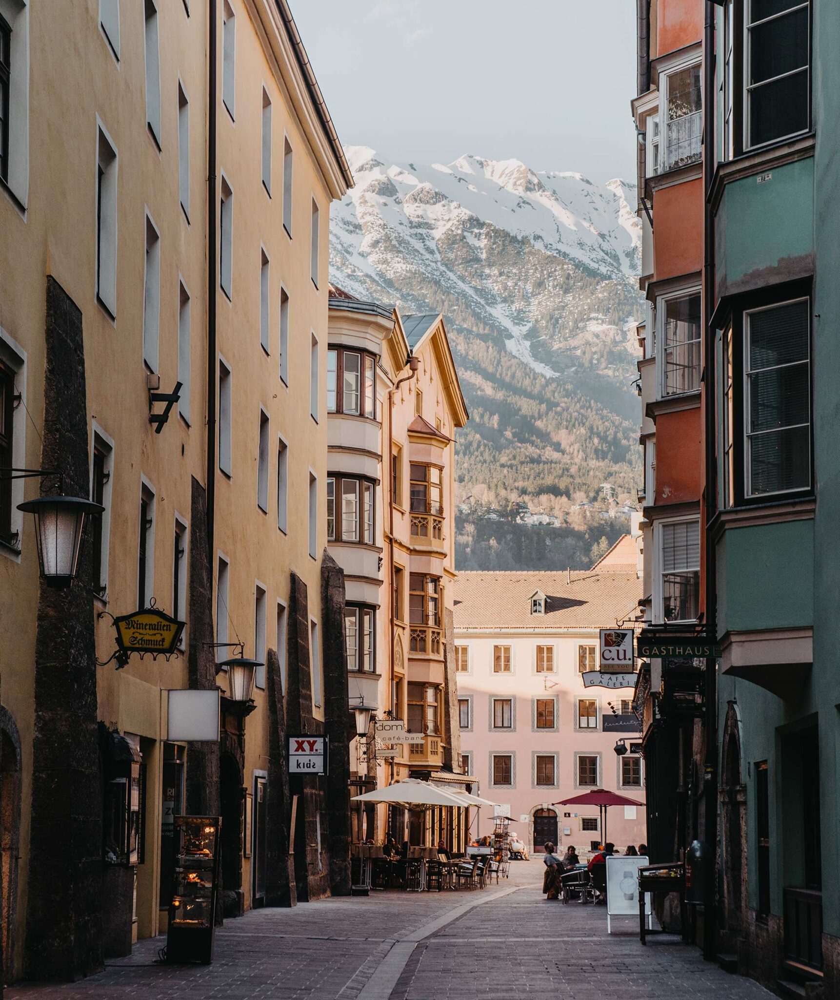Gasse in der Innsbrucker Altstadt mit bunten Häusern, Erkern und Café-Tischen, im Hintergrund die schneebedeckte Nordkette