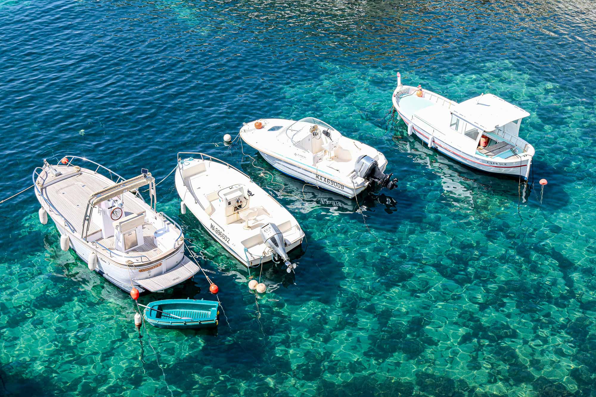 Vier weiße Motorboote und ein kleines türkisfarbenes Ruderboot ankern im glasklaren, türkisblauen Wasser an der Küste von Marseille