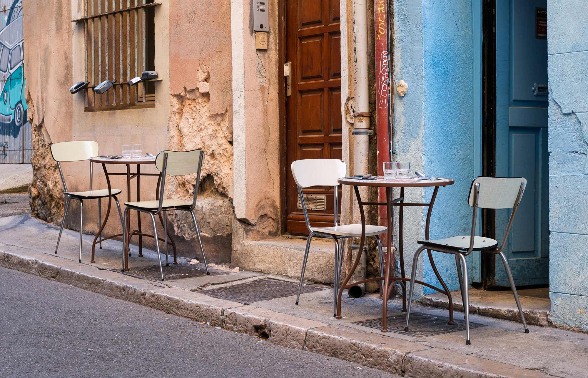 Zwei kleine Bistrotische mit Vintage-Stühlen vor verwitterter rosa und hellblauer Hausfassade in einer Gasse in Marseille
