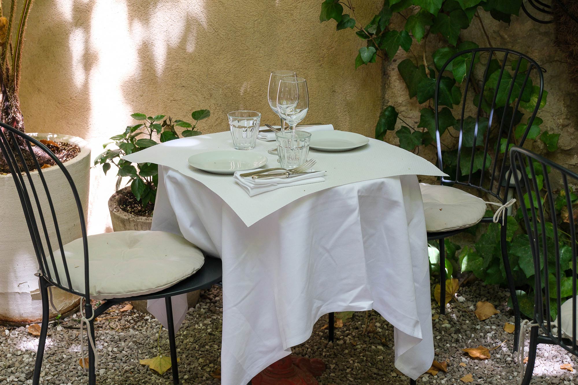 Gedeckter Tisch mit weißer Tischdecke, Weingläsern und schwarzen Metallstühlen in einem schattigen, mit Efeu bewachsenen Restaurant-Innenhof in Marseille