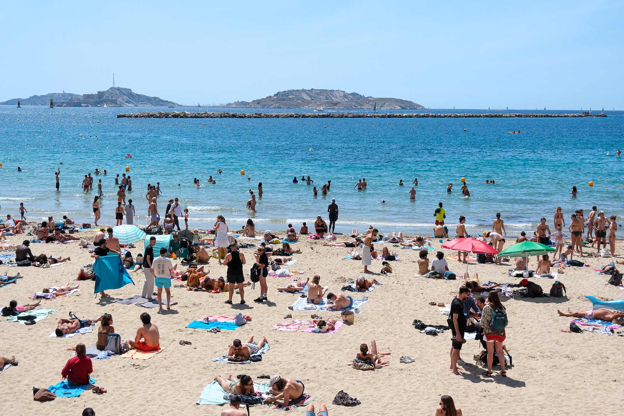 Belebter Stadtstrand in Marseille mit Badegästen, Sonnenschirmen und türkisblauem Meer, im Hintergrund die Frioul-Inseln