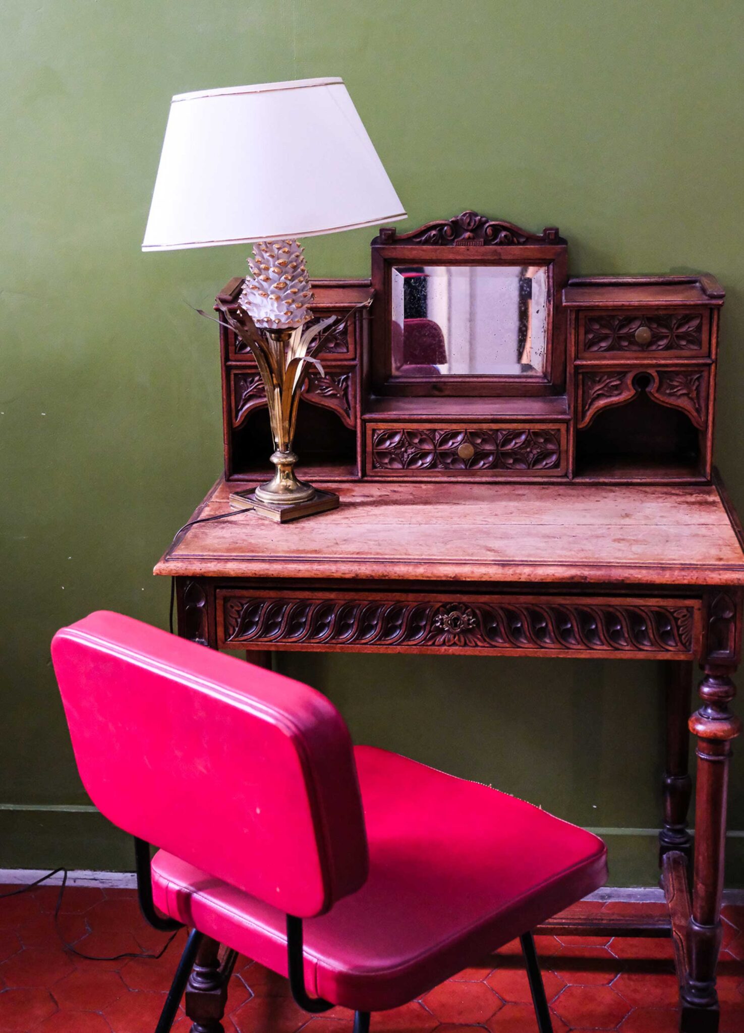 Geschnitzter Holzsekretär mit Spiegel, Tischlampe mit Messingfuß und pinkfarbenem Vintage-Stuhl vor grüner Wand in der Pension Edelweiss in Marseille