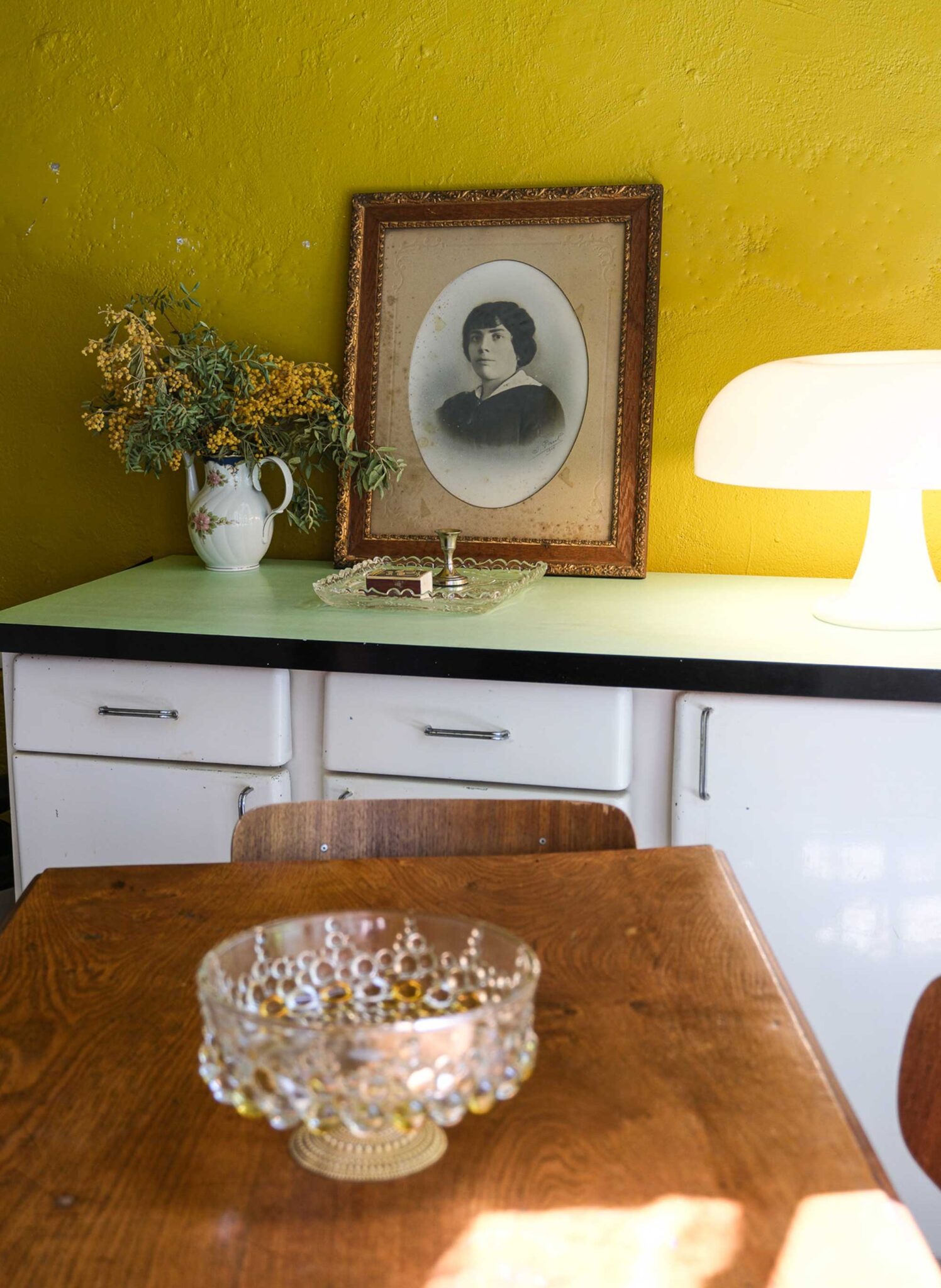 Senfgelbe Wand mit Vintage-Sideboard, altem Porträt im Goldrahmen, Mimosenstrauß und weißer Pilzlampe in der Pension Edelweiss in Marseille