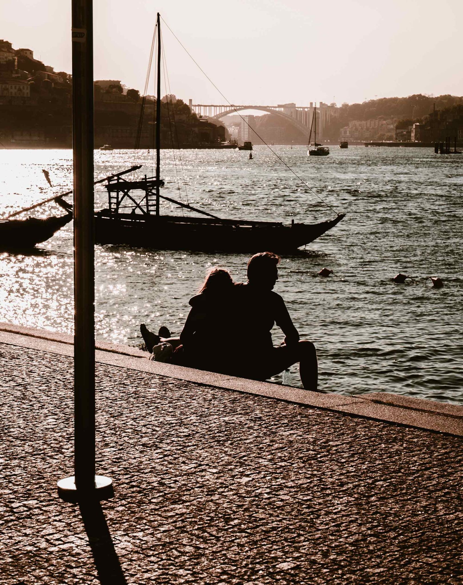 Silhouette eines Paares, das am Ufer des Douro sitzt, davor ein traditionelles Rabelo-Boot, im Gegenlicht die Arrábida-Brücke
